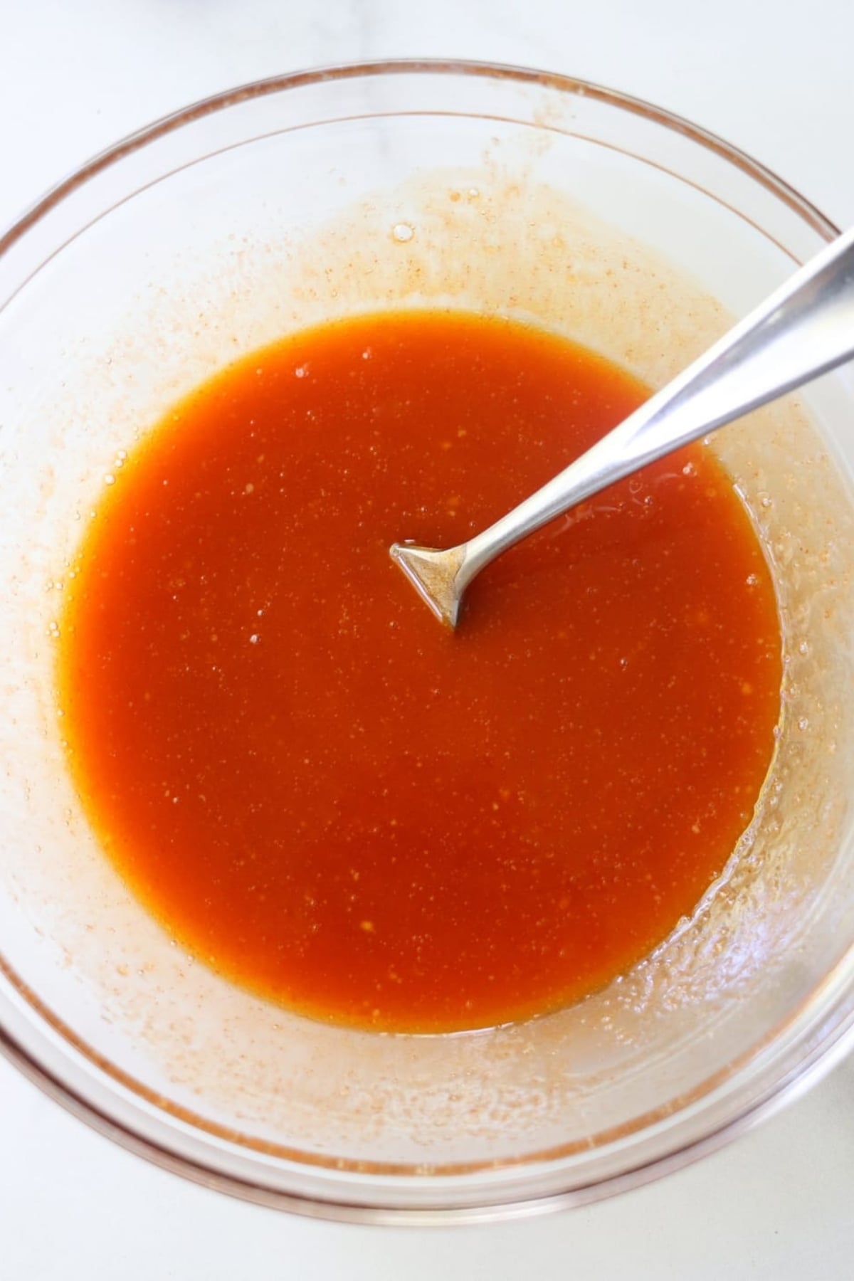 A glass bowl filled with vibrant homemade buffalo sauce, its orange-red liquid gleaming as a metal spoon rests inside.