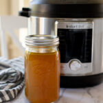 A mason jar filled with liquid is placed in front of an Instant Pot on a marble countertop, with a striped cloth nearby.