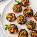 A white plate with sausage stuffed mushrooms garnished with chopped herbs and red bell peppers, placed on a light surface with a gray napkin nearby.