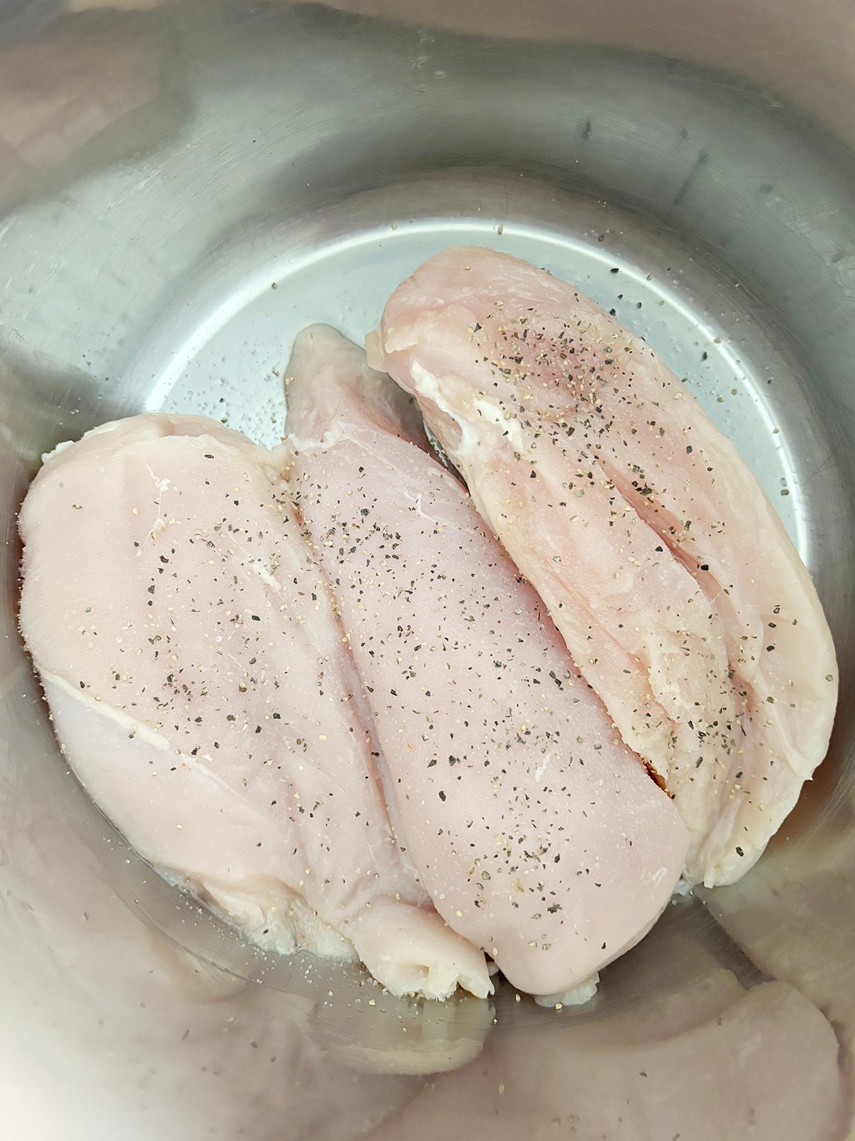 Three raw chicken breasts seasoned with salt and pepper, inside of a stainless steel pot.