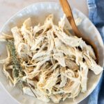 A plate of shredded chicken with a wooden spoon, garnished with a sprig of fresh thyme, on a white surface next to a blue cloth.