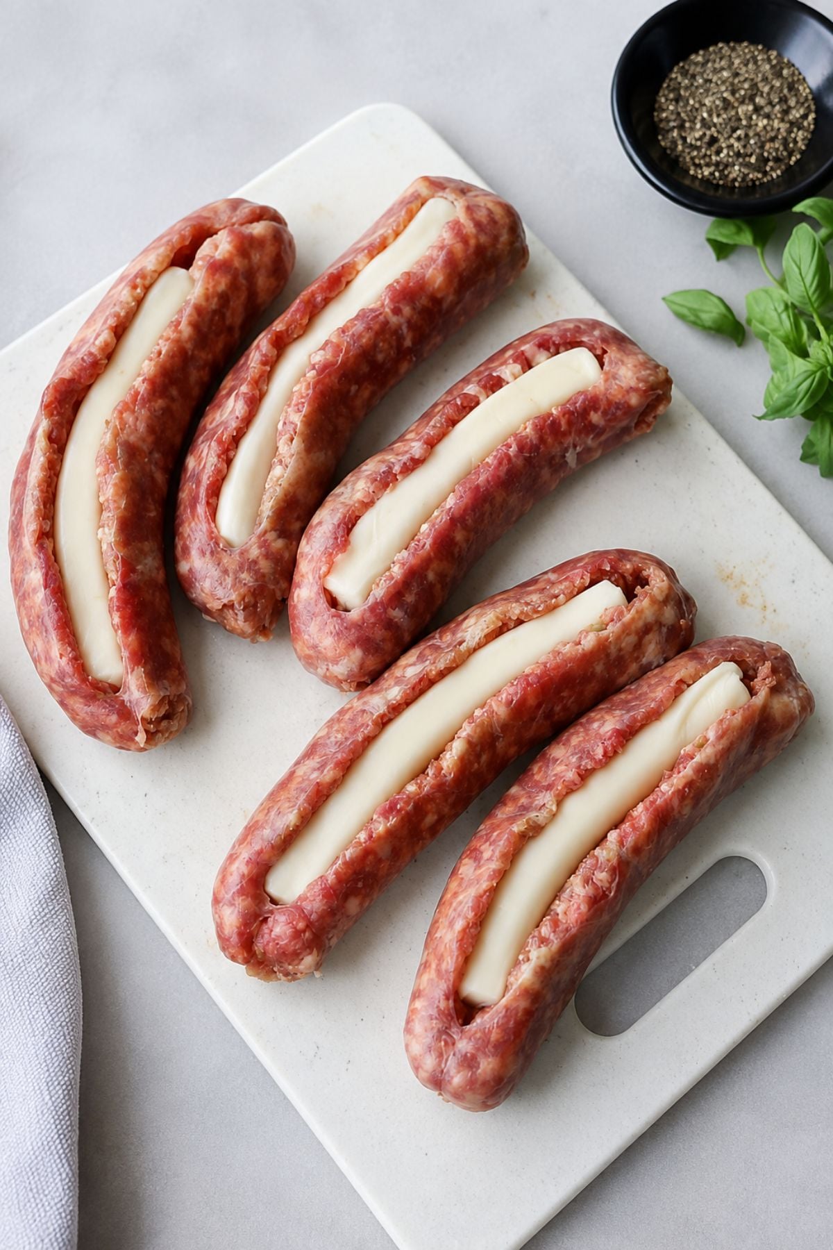 Five raw stuffed Italian sausages sliced open and filled with mozzarella cheese rest on a white cutting board, accompanied by a bowl of black pepper and fresh basil nearby.