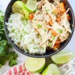A bowl of coconut lime chicken and diced vegetables next to a serving of white rice, garnished with lime wedges and fresh cilantro. Two cut limes and a red-striped cloth are placed beside the bowl.