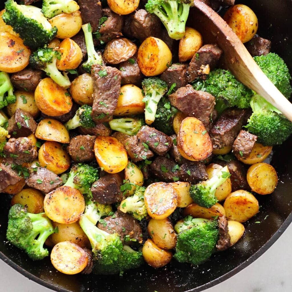 A skillet of diced beef, roasted potatoes, and broccoli, stirred with a wooden spoon.