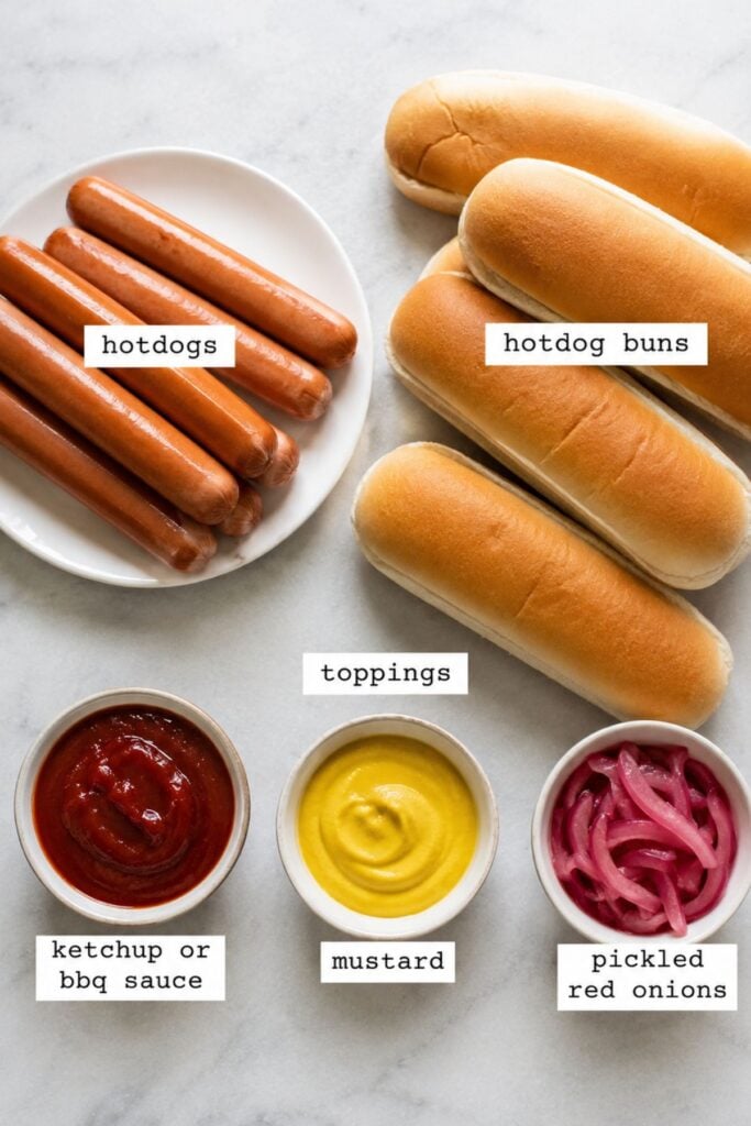 A plate of air fryer hot dogs, three hotdog buns, and small bowls of ketchup or BBQ sauce, mustard, and pickled red onions are arranged on a white surface.