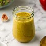 A glass jar filled with creamy, yellow, apple cider vinaigrette salad dressing, placed on a marble surface. Nearby are a spoon, a garlic clove, black pepper, and a red apple. A salad bowl is visible in the background.