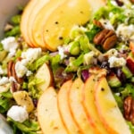 Brussel sprouts salad with apple slices, pecans, feta cheese, onions, and greens in a white bowl.