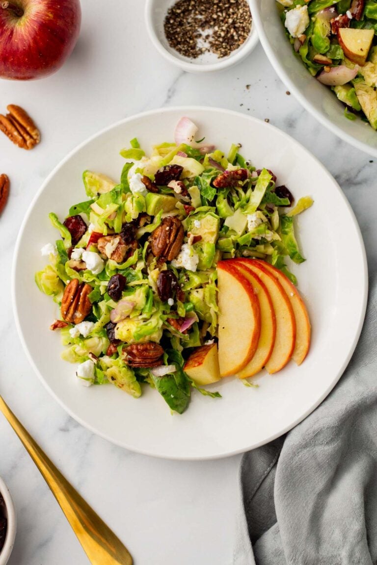 Plate of Brussel sprouts salad with avocado, pecans, feta, cranberries, and sliced apples on a white marble surface. A gold fork, salt dish, and apple are nearby.