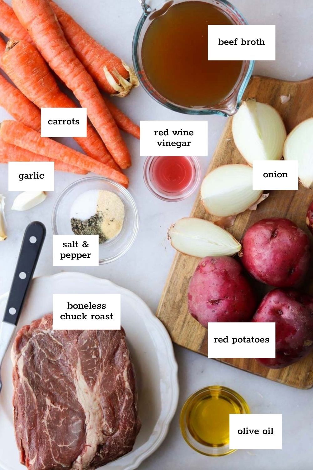 Ingredients for a pot roast recipe laid out on a white surface, including carrots, garlic, onion, red potatoes, boneless chuck roast, beef broth, vinegar, olive oil, and seasonings.
