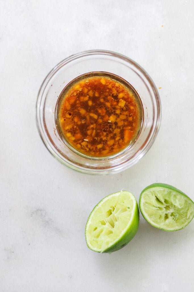 A bowl of soy ginger marinade beside two squeezed lime halves on a white surface.