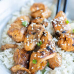 A bowl of cooked white rice topped with teriyaki salmon pieces, garnished with sesame seeds and chopped green onions, with chopsticks on the side.