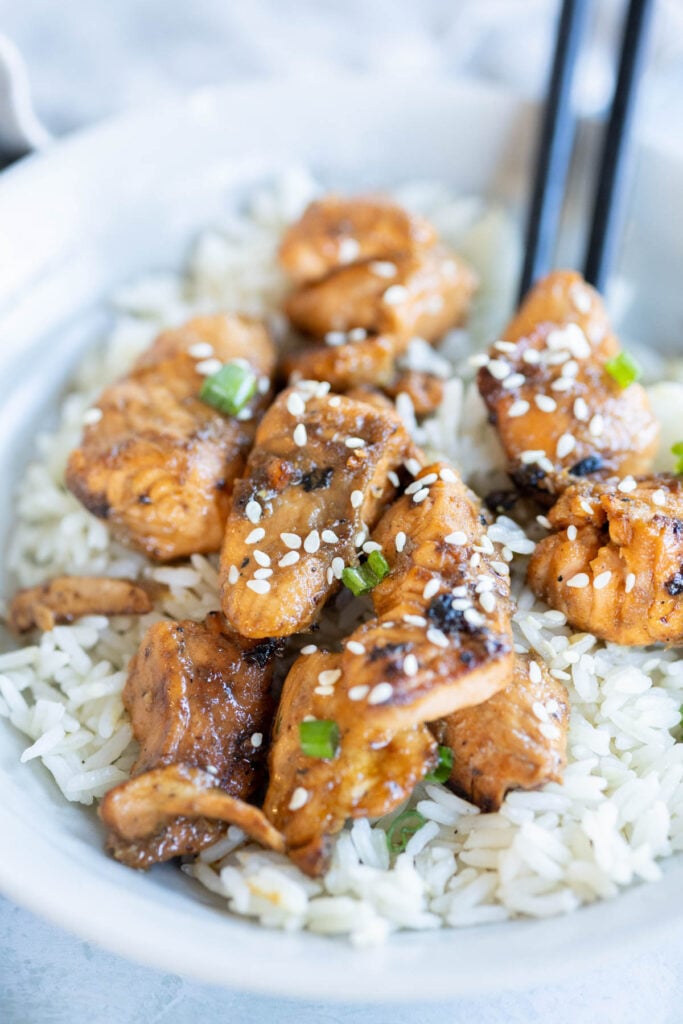 A bowl of cooked white rice topped with teriyaki salmon pieces, garnished with sesame seeds and chopped green onions, with chopsticks on the side.