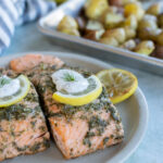 Two marinated salmon fillets baked with dill and garnished with lemon slices and sauce on a plate. A striped cloth and a tray of roasted potatoes are in the background.