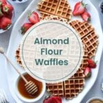 A plate of Paleo waffles made with almond flour, topped with strawberries and honey, is surrounded by bowls of blueberries, juicy strawberries, and a steaming cup of coffee. Perfect for your own waffle bar setup at home.