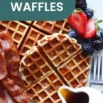 A plate of paleo waffles made with almond flour, served alongside crispy bacon and fresh berries. A small pitcher of syrup waits to drizzle over this delightful breakfast. Text on image: "Waffle Bar - Almond Flour Goodness.