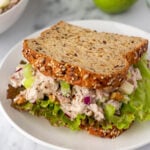 A chicken salad sandwich with lettuce on multigrain bread is on a white plate. Green apples and a bowl of chicken salad are in the background.