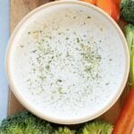 A bowl of healthy ranch dressing garnished with herbs, surrounded by red bell pepper slices and broccoli on a wooden surface.