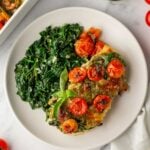 Plate of lasagna with cherry tomatoes and basil, served with sautéed spinach. Jar of pesto and fresh tomatoes in the background.