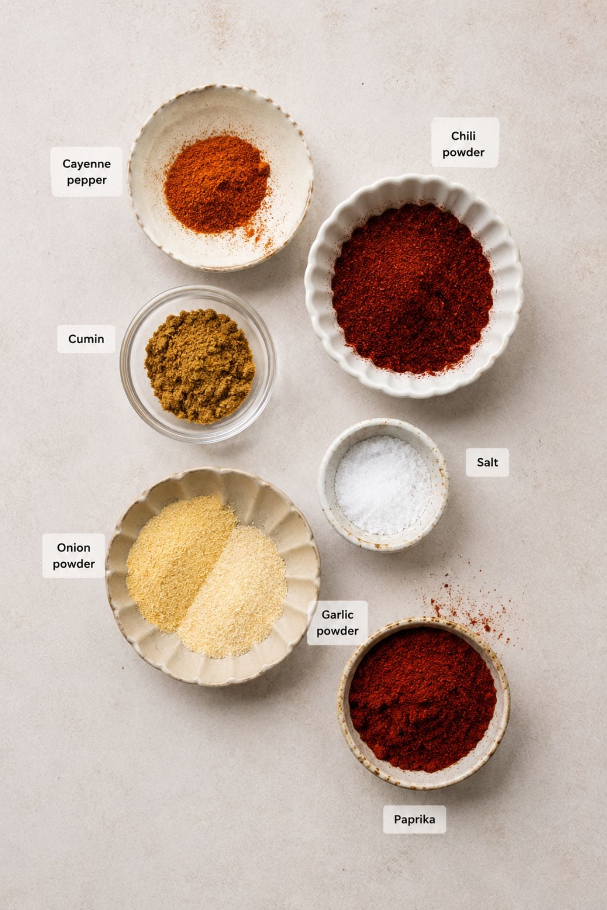 Six small bowls containing ground spices—cayenne pepper, chili powder, cumin, salt, onion powder, garlic powder, and paprika—arranged on a light surface with each labeled. These classic ingredients come together perfectly to create a homemade fajita seasoning blend.