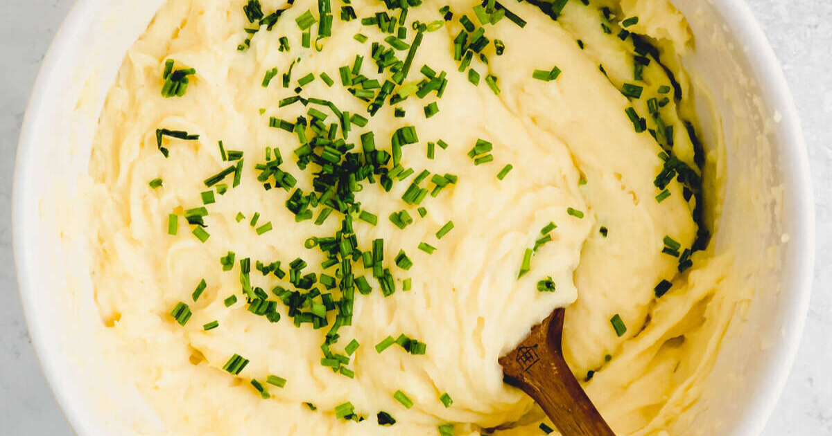 A bowl of mashed potatoes topped with chopped chives, with a wooden spoon resting in the mixture.