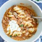 A bowl of lasagna soup with a tomato-based broth, spiral noodles, ground meat, and topped with a dollop of ricotta cheese. A spoon is in the bowl. Chopped herbs are visible in a dish nearby.