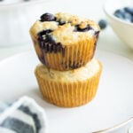 Two blueberry muffins stacked on a white plate, surrounded by a striped cloth and a bowl of blueberries.