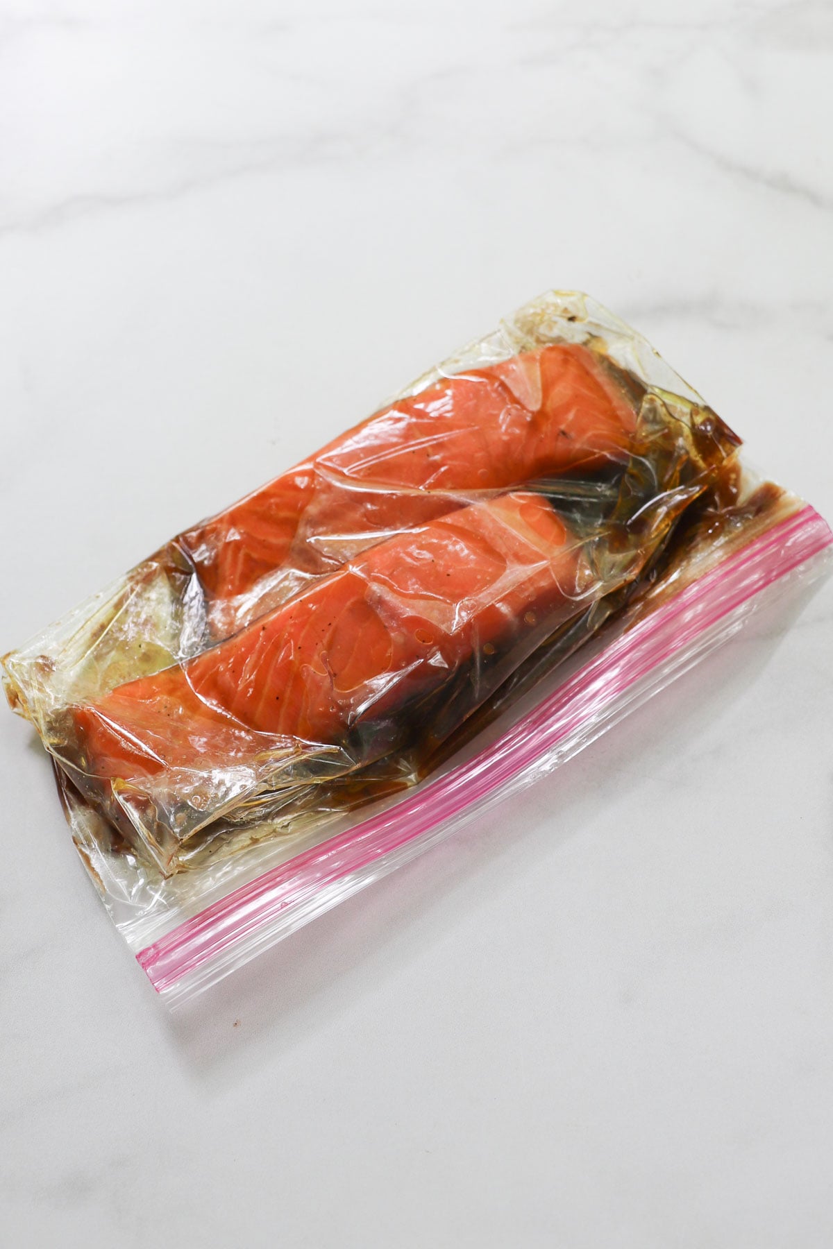 Two raw salmon fillets marinating in a clear plastic ziplock bag on a white surface.