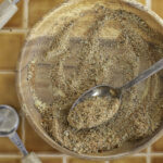 Wooden bowl with ground spices and a metal spoon on a tiled surface, surrounded by measuring spoons.