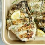 Ranch chicken breast with grill marks. Lemon wedges on a beige tray, garnished with herbs.