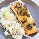 Cooked salmon fillet topped with spicy green chiles, served with white rice and lime wedges on a gray plate.