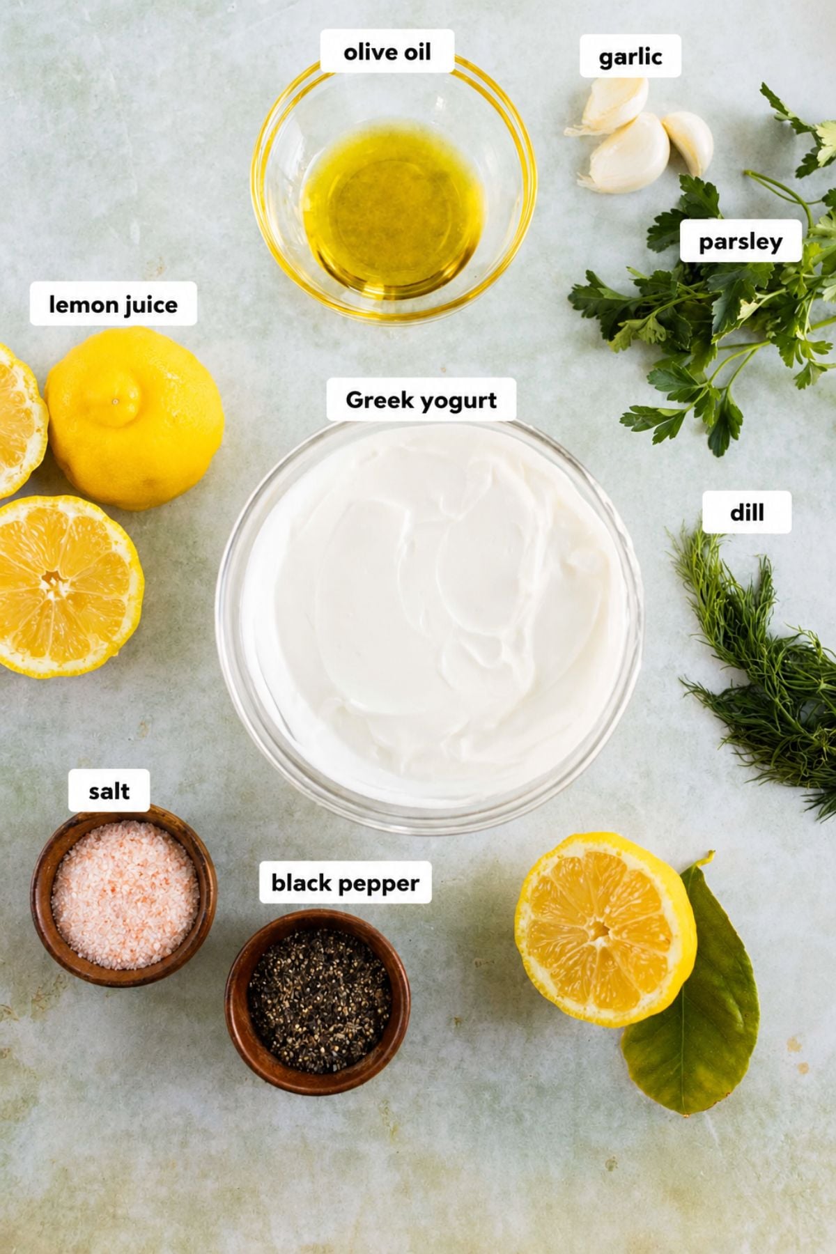 Ingredients for a dipping sauce including yogurt, lemon halves, olive oil, garlic cloves, parsley, dill, pink salt, and black pepper, arranged on a light surface.