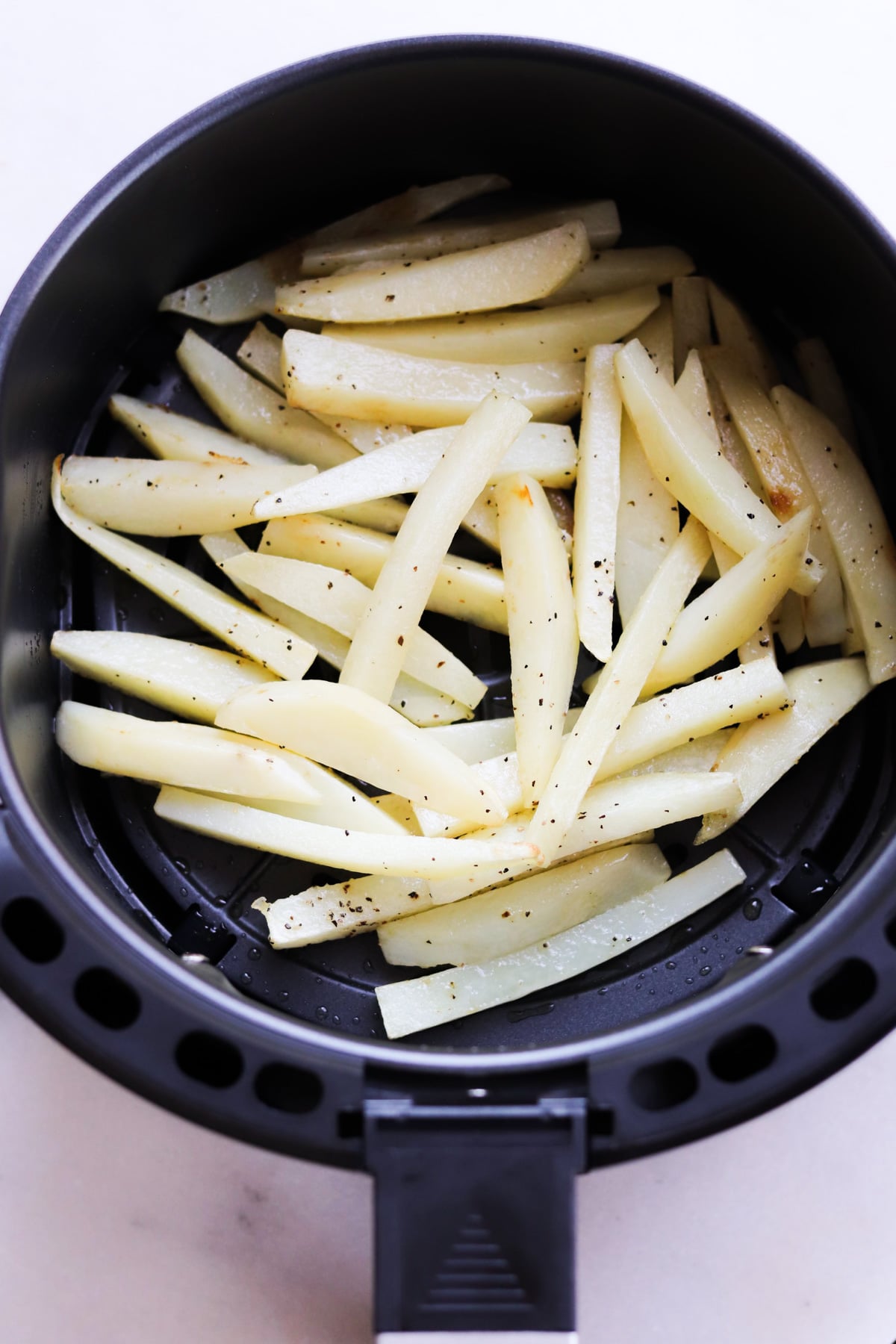 Raw potato strips seasoned with salt and pepper are arranged in an air fryer basket, ready to be cooked.