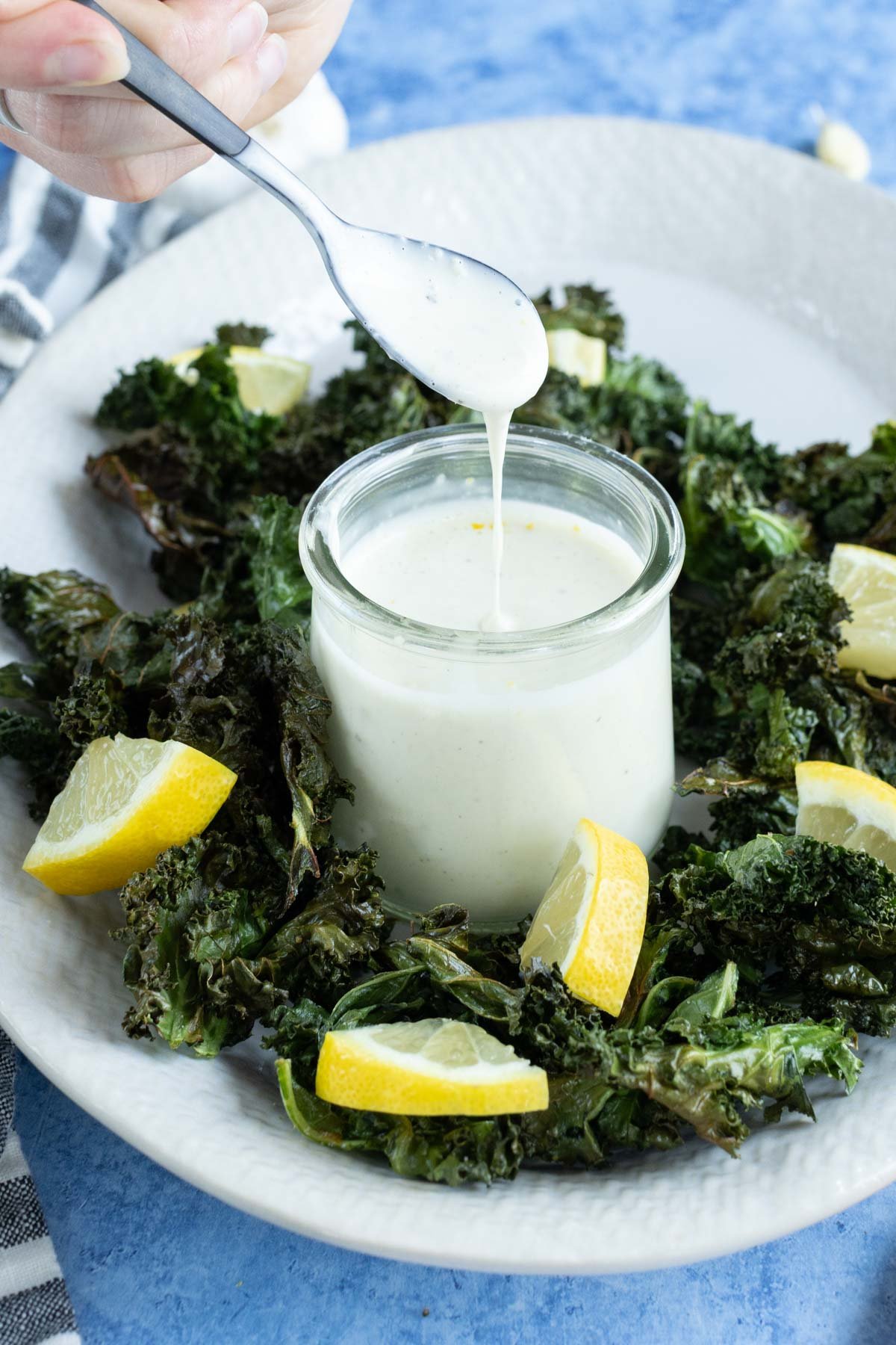 A small jar of creamy dressing is surrounded by kale chips and lemon wedges on a white plate, with a hand holding a spoon above the dressing.