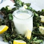 A person's hand dips a kale chip into a small jar of creamy white sauce. The plate is garnished with crispy kale chips and lemon wedges.