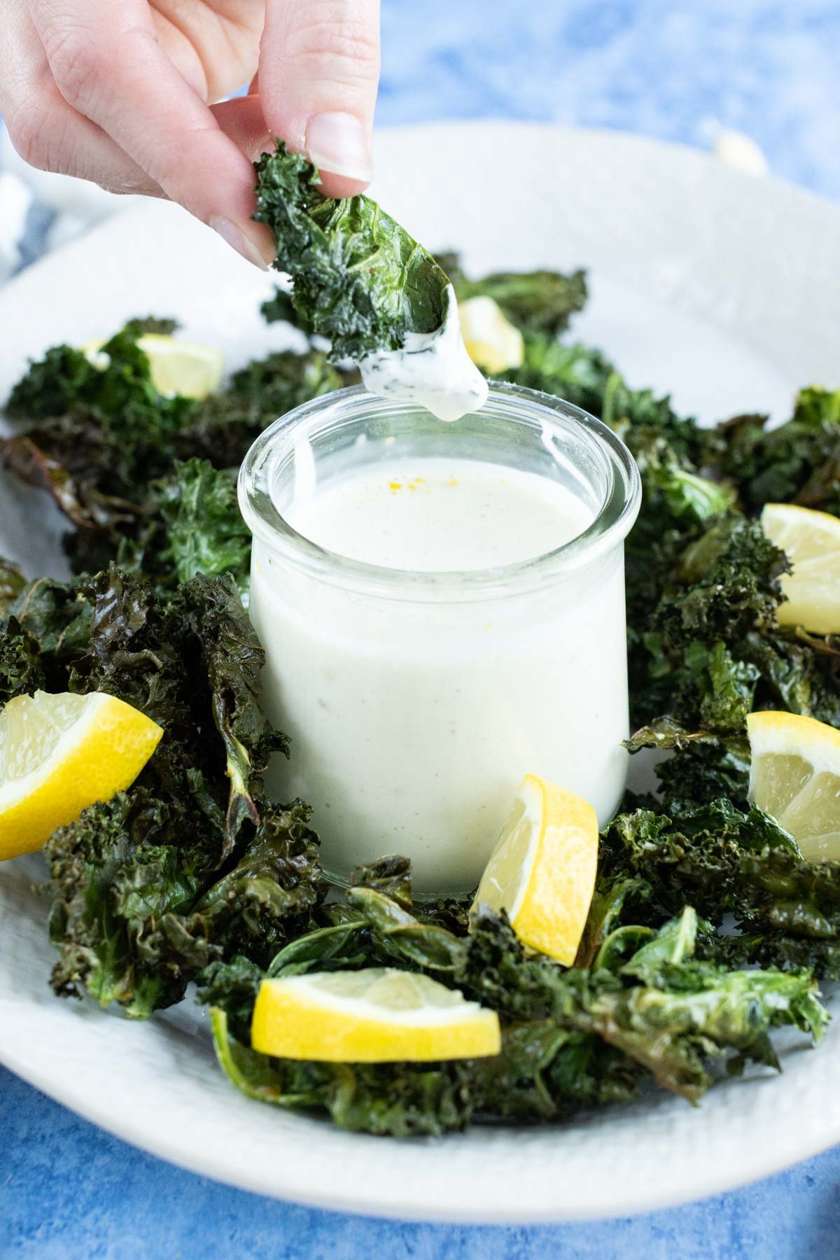 A person's hand dips a kale chip into a small jar of creamy white sauce. The plate is garnished with crispy kale chips and lemon wedges.