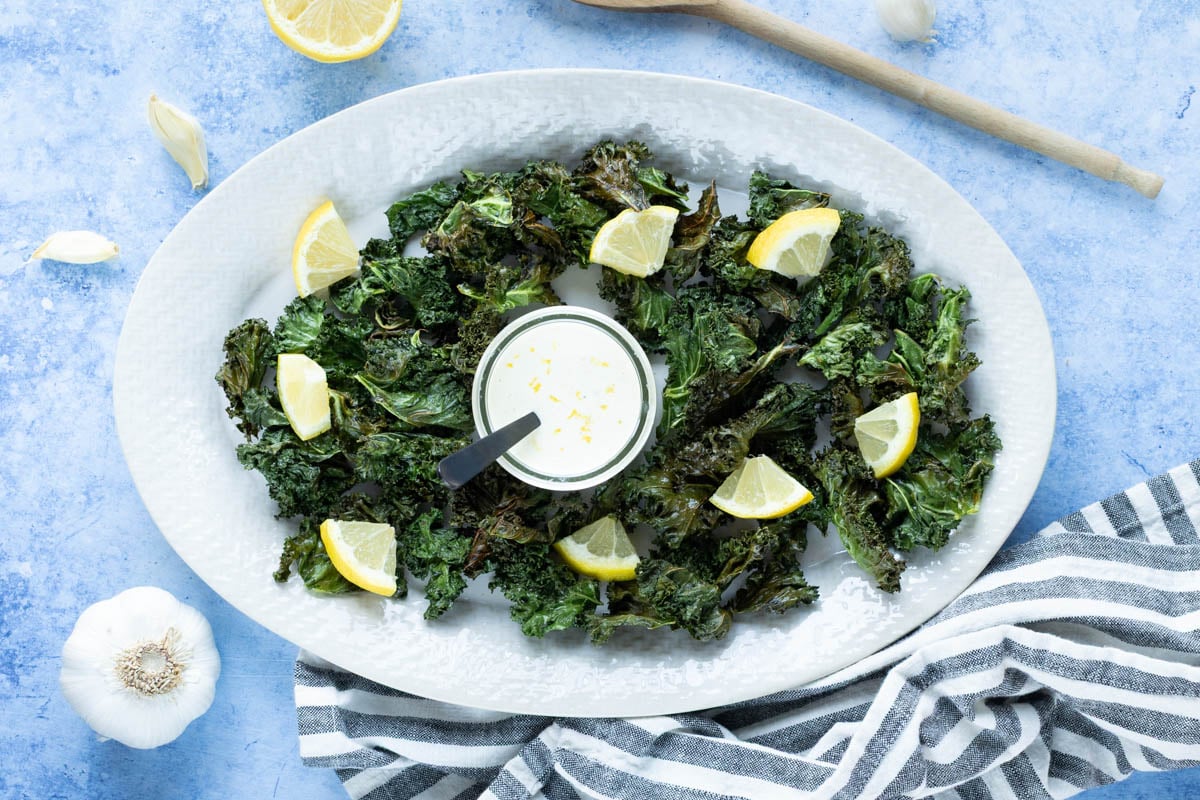 Kale chips and lemon wedges arranged on a large white plate with a small bowl of dip in the center, set on a blue surface with garlic, a striped cloth, and a wooden spoon nearby.