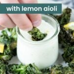 A hand dips air fryer kale into a jar of lemon aioli. The background features more crispy kale pieces and tart lemon wedges, adding a vibrant touch. Text on the image proudly declares "Air Fryer Kale with Lemon Aioli.