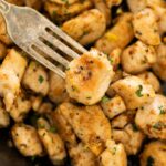A fork holds a piece of cooked, seasoned chicken above a pan filled with similar pieces. The chicken is lightly browned with herbs sprinkled on top.