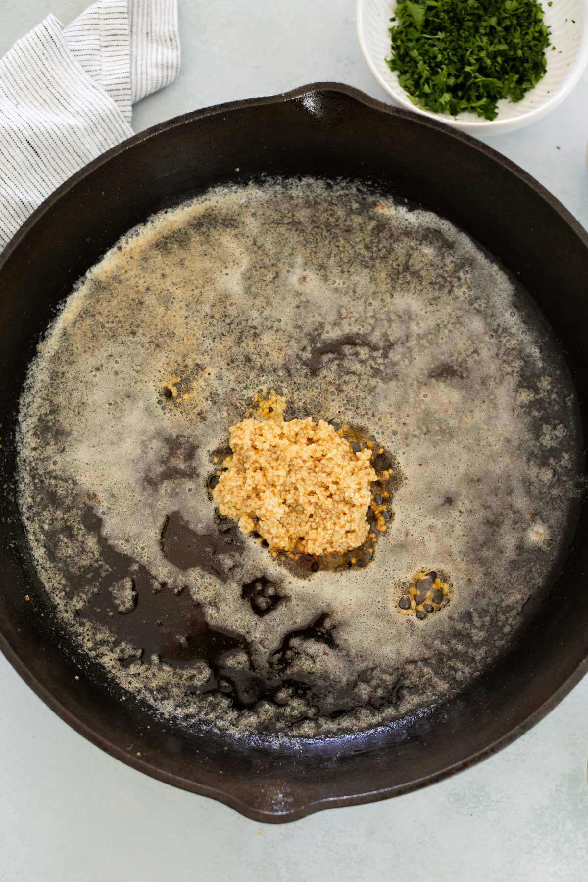 A cast iron skillet with bubbling butter and minced garlic in the center. A bowl of chopped herbs is nearby.