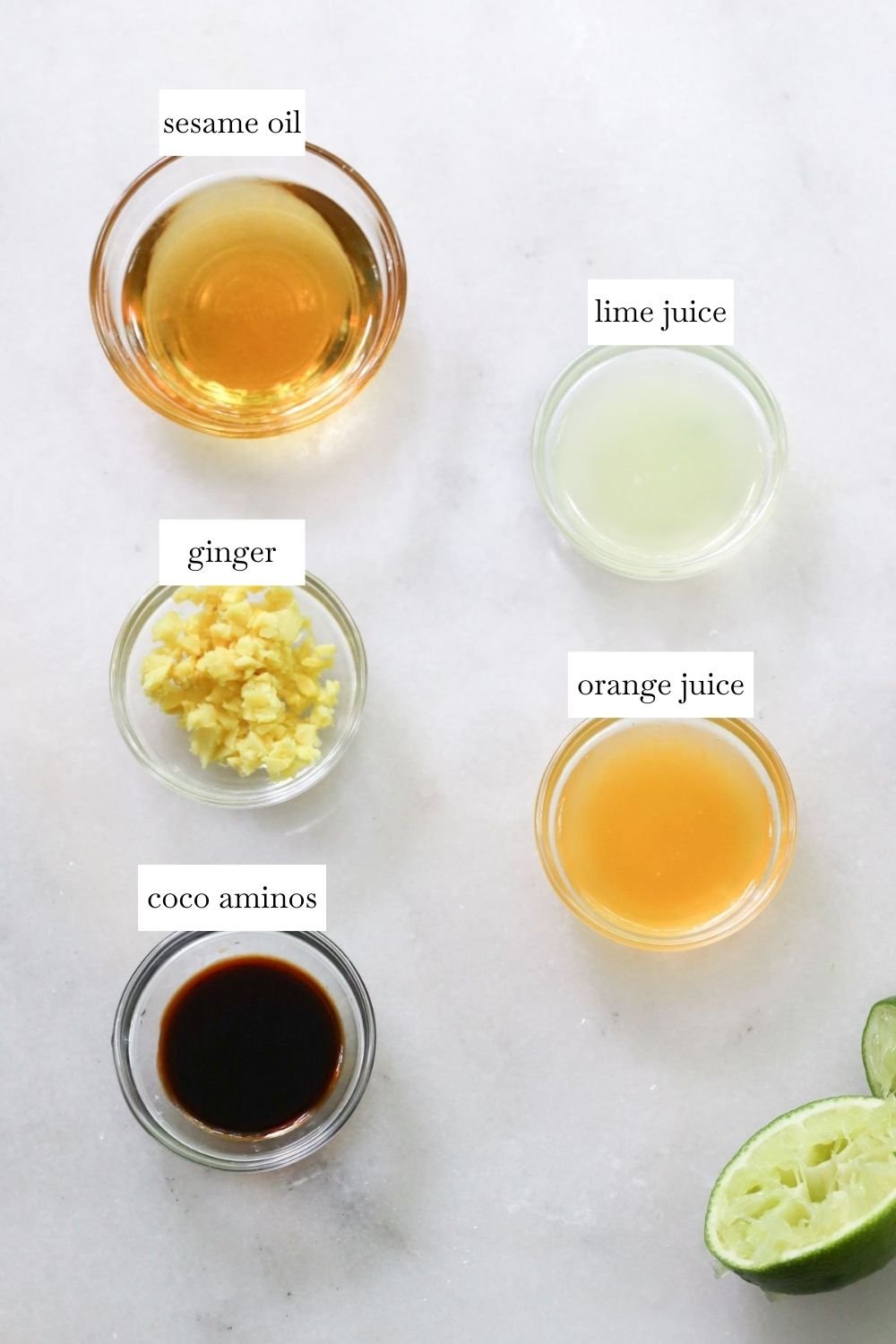 Five labeled ingredients on a white surface: sesame oil, lime juice, grated ginger, orange juice, and coco aminos, with a sliced lime.