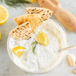 A bowl of Greek yogurt sauce garnished with dill and lemon wedges, with grilled pita triangles. Lemons, a garlic clove, and a wooden juicer are nearby.