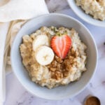 A bowl of Instant Pot oatmeal topped with banana slices and a halved strawberry, sprinkled with cinnamon. Nearby are a spoon, a cloth, a bowl of blueberries, and a strawberry on a marble surface.