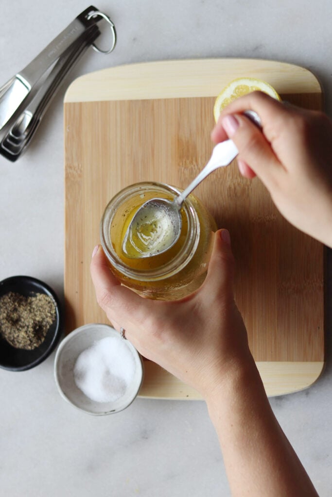 A person stirs a mixture in a jar with a spoon, above a wooden cutting board. Sliced lemon, measuring spoons, and bowls of salt and pepper are nearby.