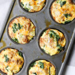 A muffin tray holds six baked egg cups with spinach and melted cheese, placed on a white cloth.