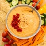 A bowl of red pepper hummus topped with diced peppers, surrounded by sliced cucumbers, carrots, yellow bell peppers, grape tomatoes, and crackers on a wooden board.