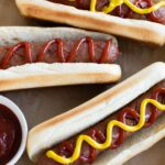 Hot dogs topped with ketchup and mustard in buns, accompanied by small bowls of ketchup and mustard.