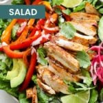 A chicken fajita salad with tender dutch oven chicken slices, bell peppers, avocado, pickled onions, lettuce, lime wedges, and cilantro in a bowl.