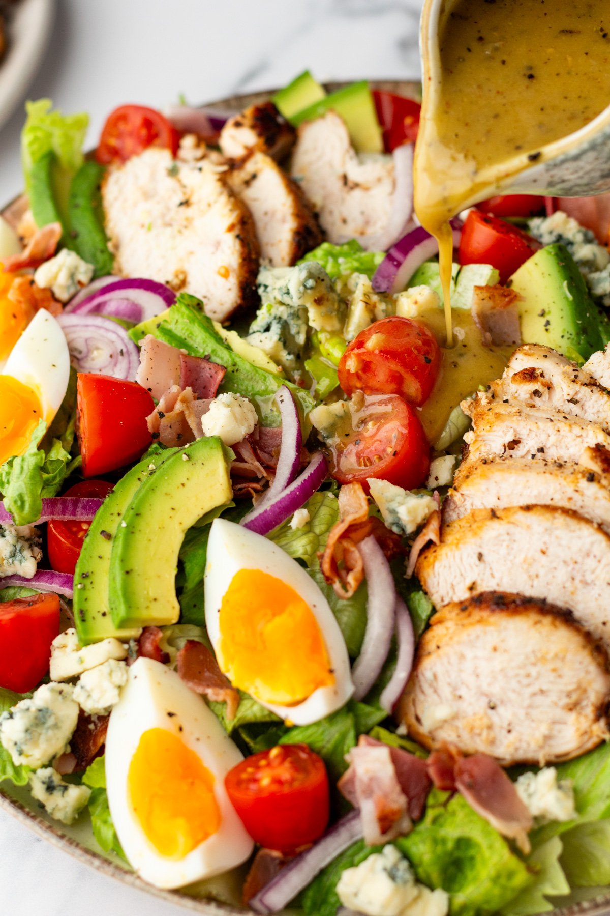 A salad with sliced grilled chicken, avocado, cherry tomatoes, red onion, blue cheese, hard-boiled egg, and lettuce, with dressing being poured on top.