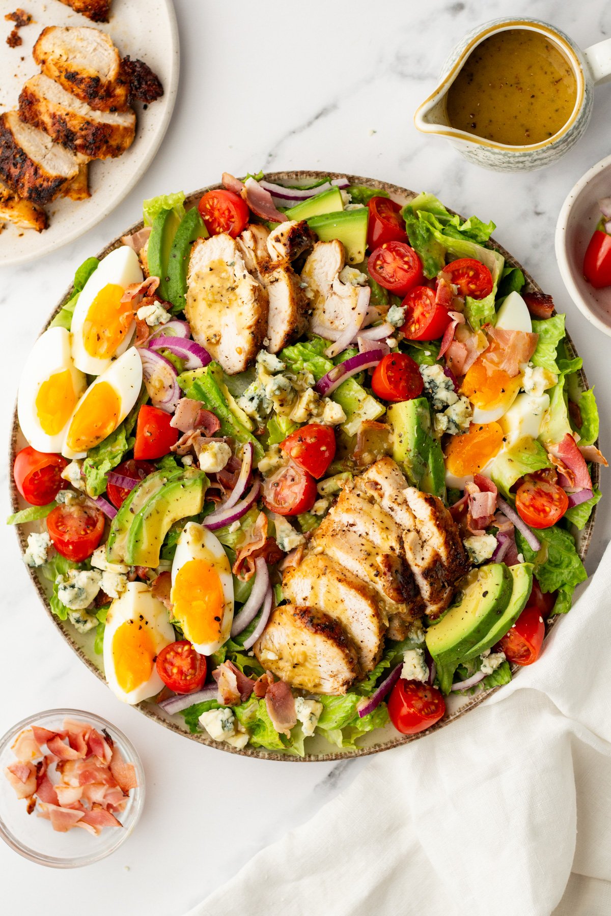 A salad with sliced grilled chicken, hard-boiled eggs, avocado, cherry tomatoes, red onion, bacon, and blue cheese on a bed of greens, with dressing and ingredients on the side.