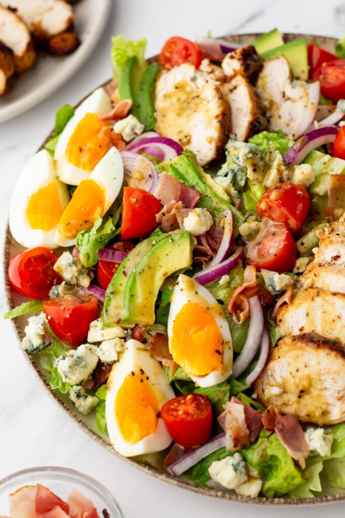 A salad bowl with sliced grilled chicken, avocado, cherry tomatoes, red onion, blue cheese, prosciutto, and halved soft-boiled eggs on a bed of lettuce.